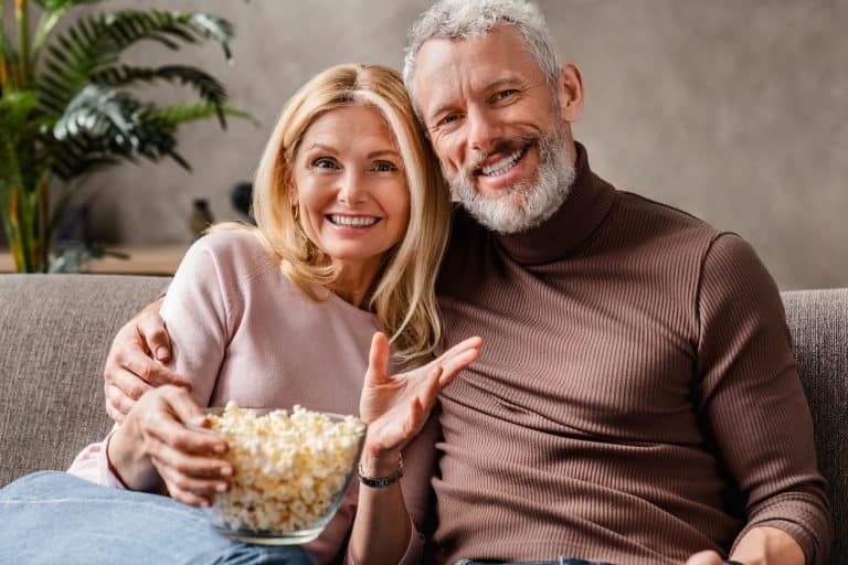 Happy middle aged couple, wife and husband watching tv and eating popcorn