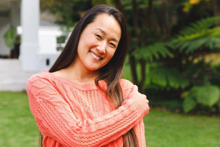 Portrait of happy asian woman smiling in garden outside family home wearing pink sweater